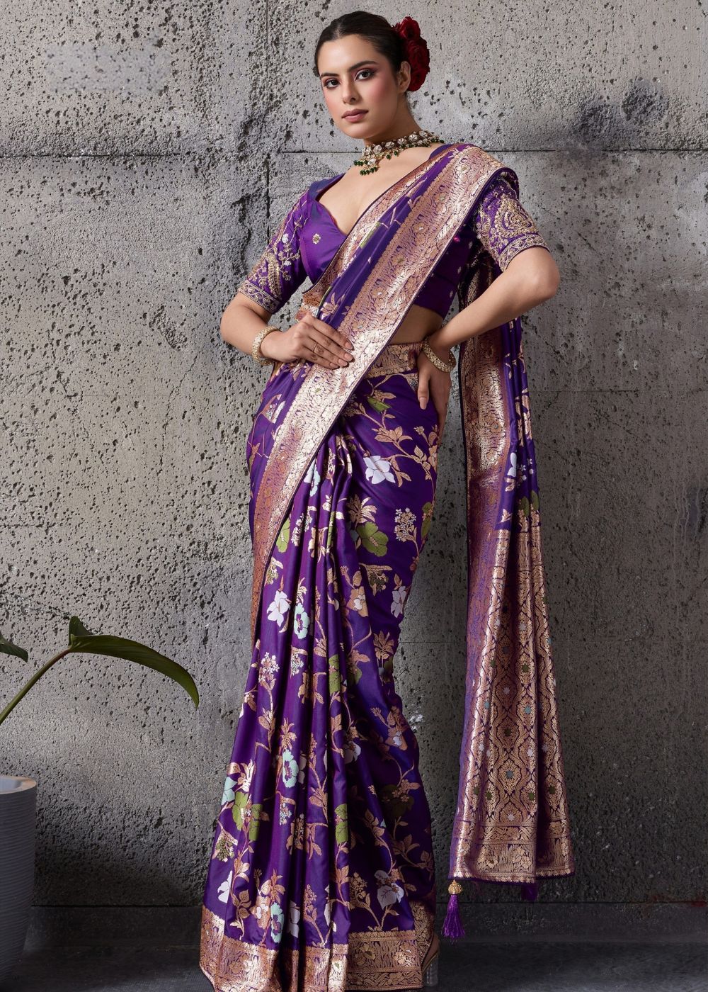Woman wearing a purple saree with floral patterns against a gray concrete wall.